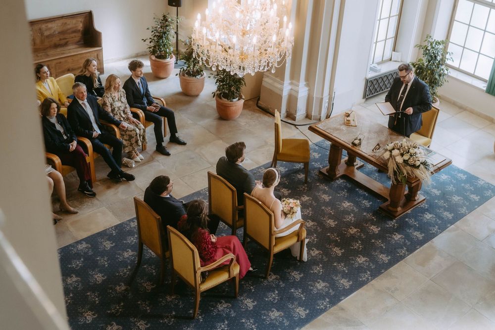 A small indoor ceremony takes place in an elegant room with a crystal chandelier and wooden chairs on a blue patterned carpet.