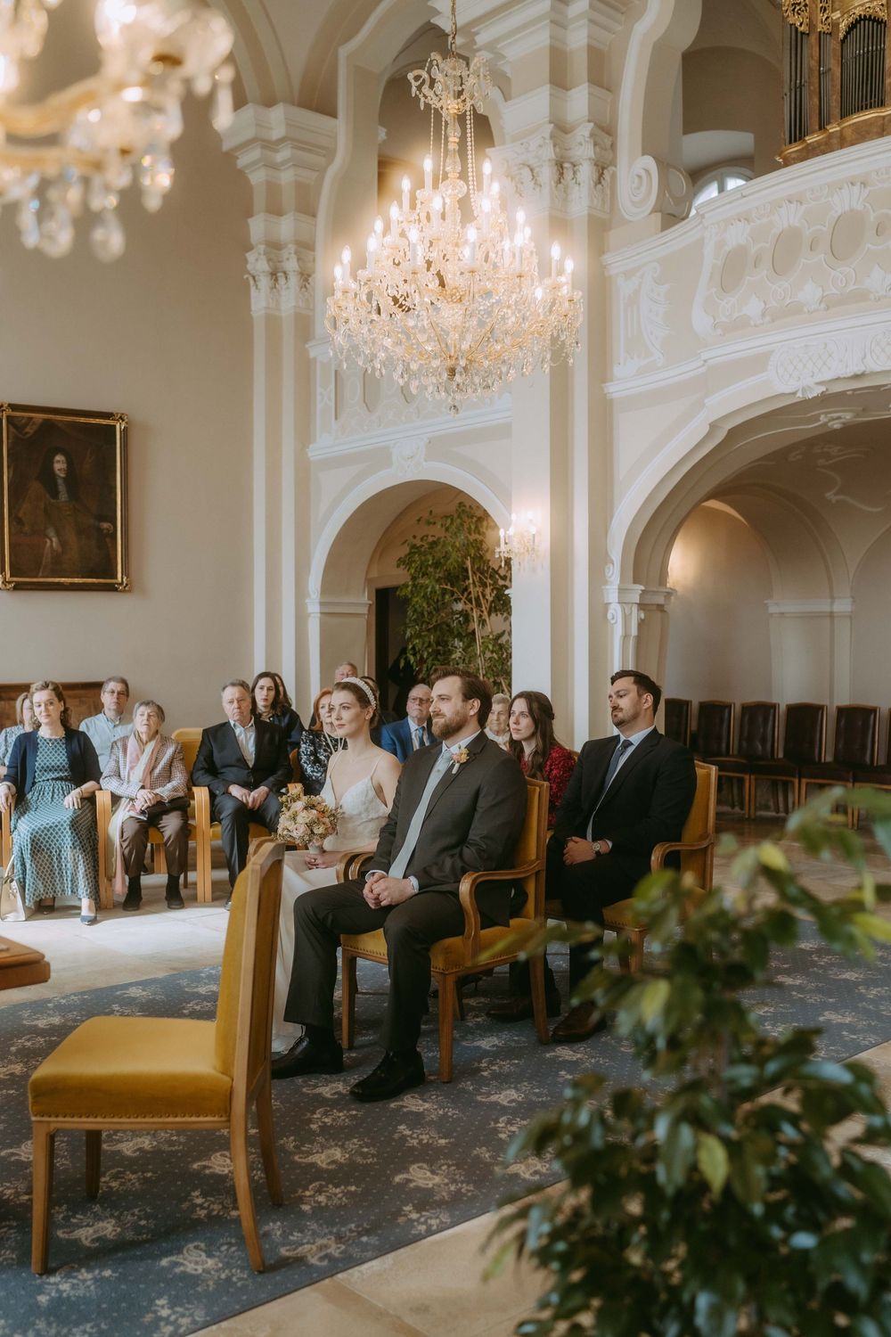 Elegant wedding ceremony in an ornate room with crystal chandeliers, white columns, and guests seated in wooden chairs.