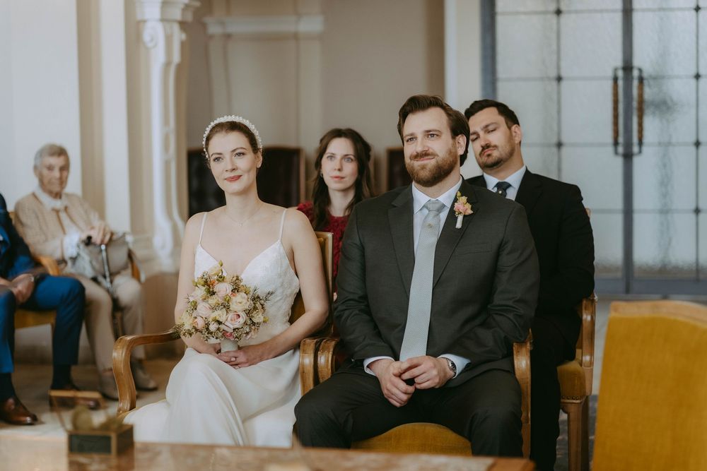 A couple sits together during an intimate indoor wedding ceremony with white columns and yellow chairs in the background.