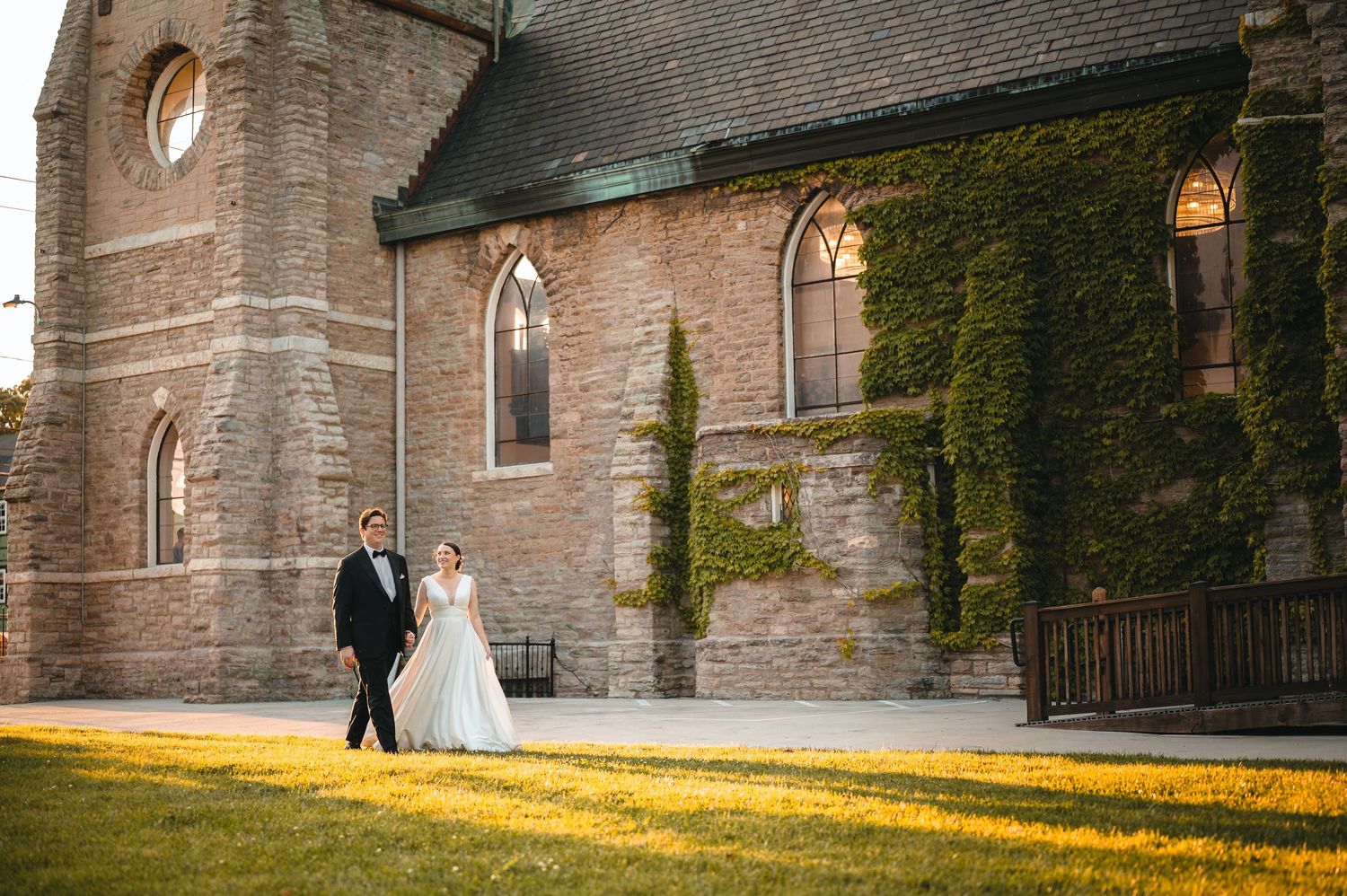 A couple stands outside an ivy-covered stone church with Gothic windows during golden hour sunlight.