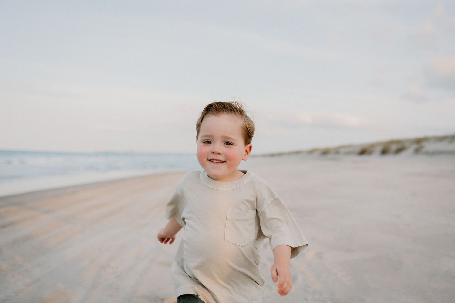 A young child runs and plays joyfully along a sandy beach during a cloudy day.