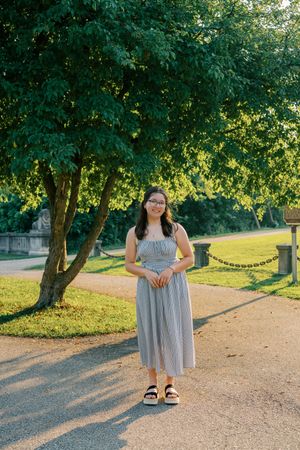 A person in a striped gray sundress stands on a path under a leafy green tree on a sunny day.