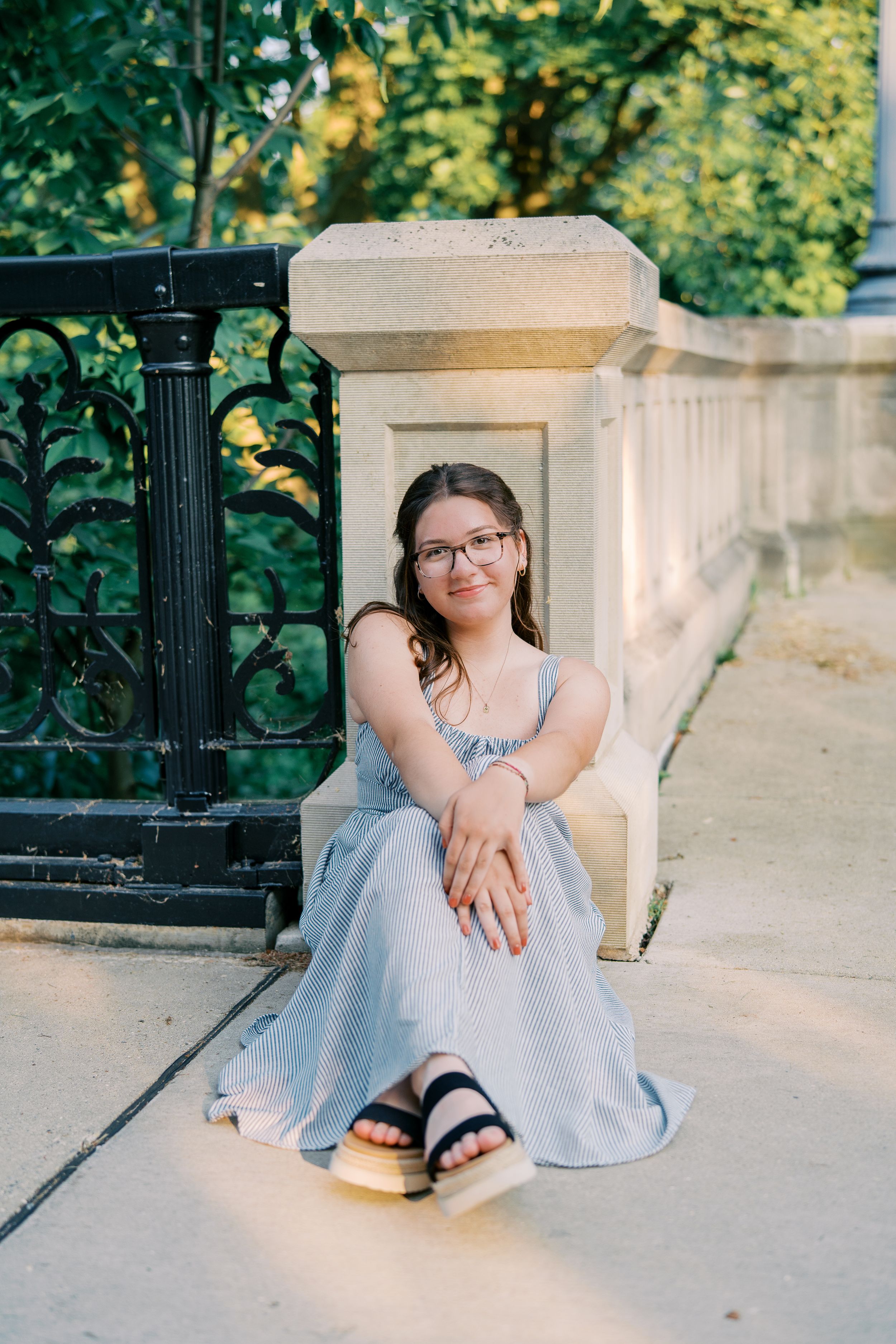 A person in a gray dress sits on stone steps near an ornate black iron railing.
