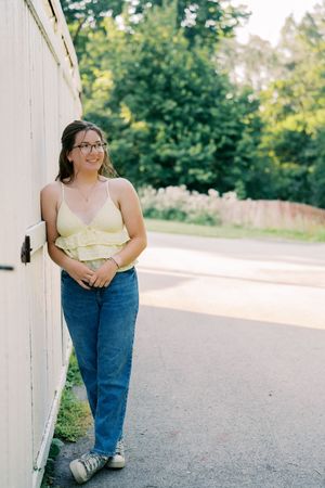 Person in yellow top and jeans poses casually against white wall outdoors.