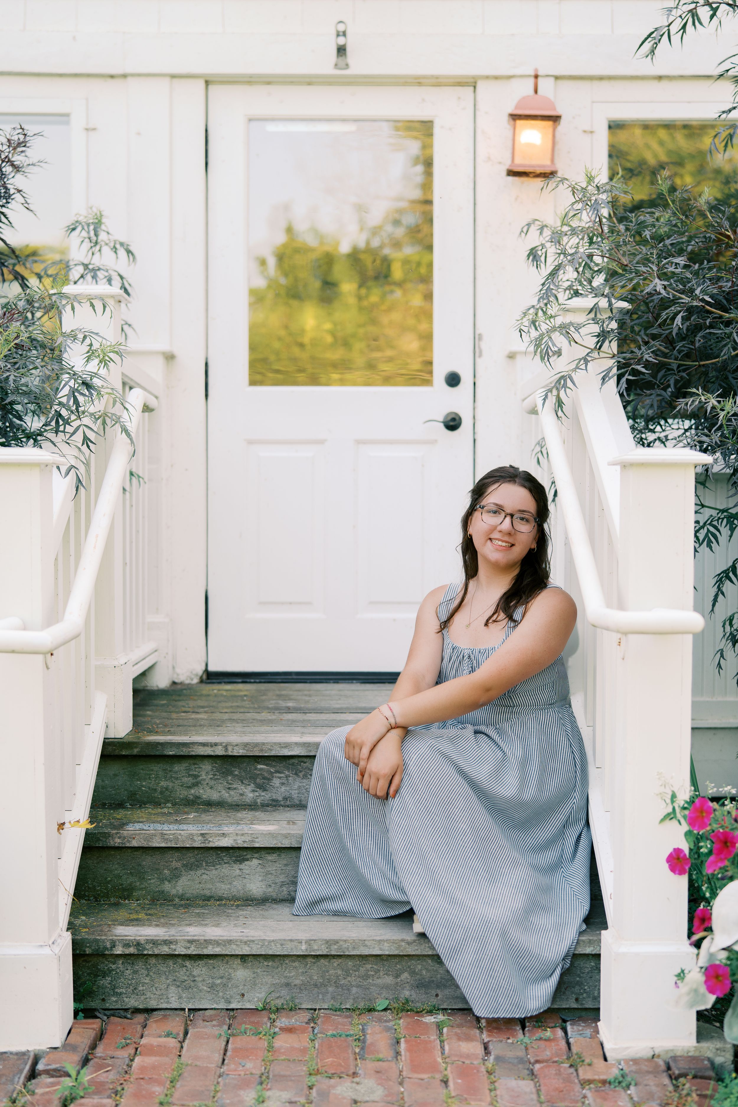 Someone sits on brick steps wearing a gray dress near white pillars and flowering bushes.