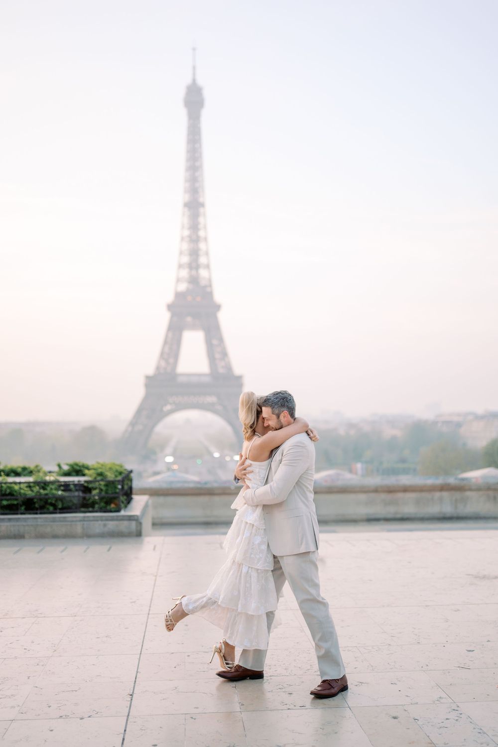 A romantic couple in white attire shares an intimate moment with the Eiffel Tower visible in the misty background.