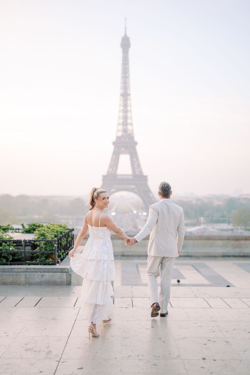 A couple in white attire poses for romantic wedding photos with the iconic Eiffel Tower visible in the misty Paris backdrop.