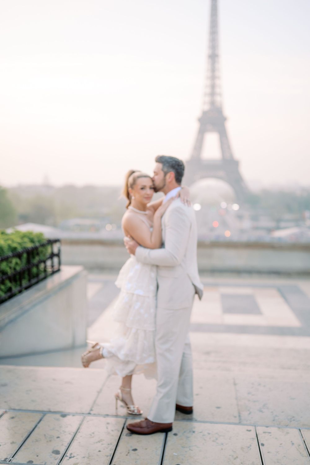 A romantic couple in white outfits embraces near the Eiffel Tower in Paris during a dreamy photoshoot at sunset.