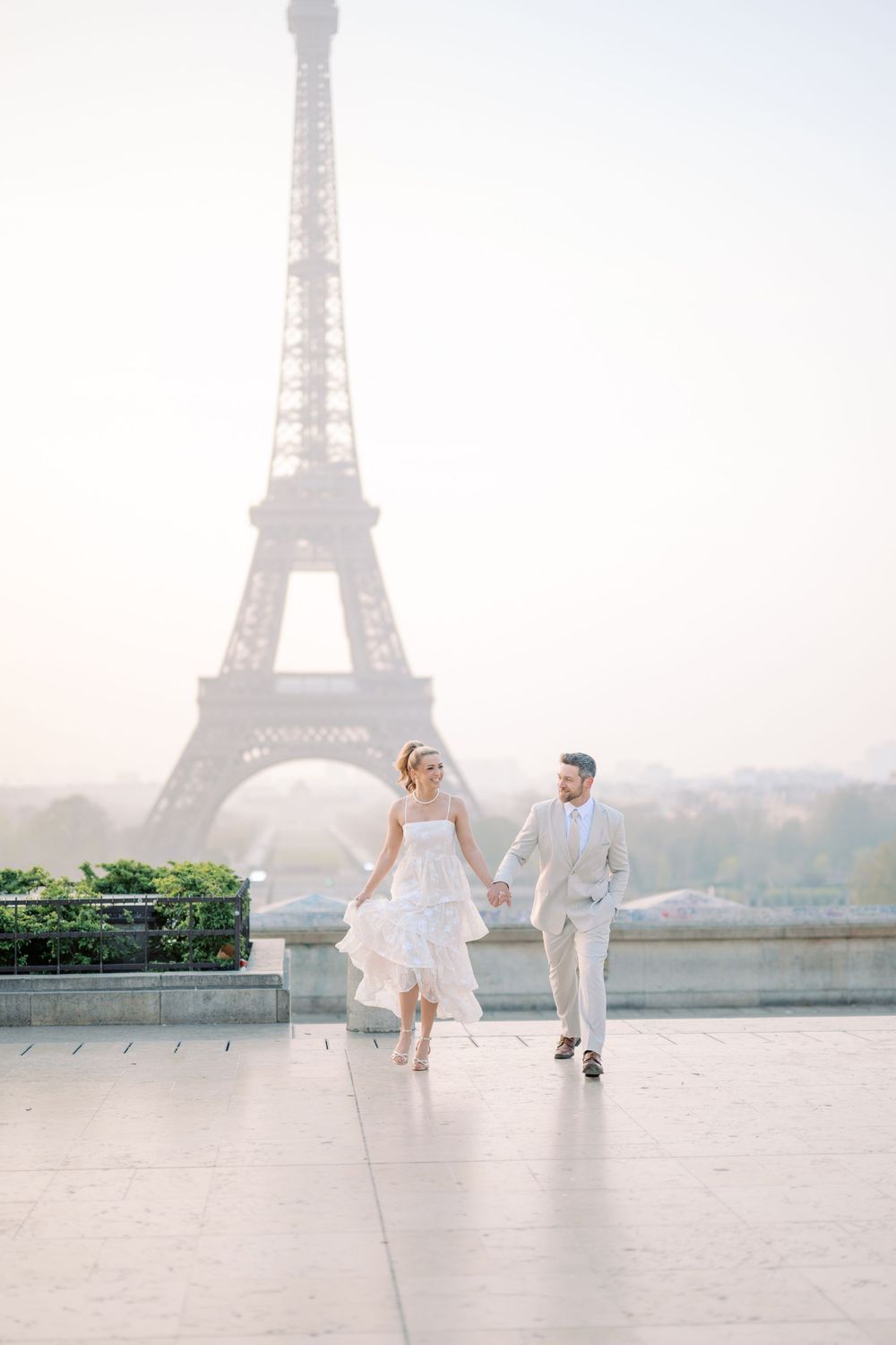 A romantic couple in wedding attire runs joyfully at Trocadero plaza with the Eiffel Tower in the misty background.