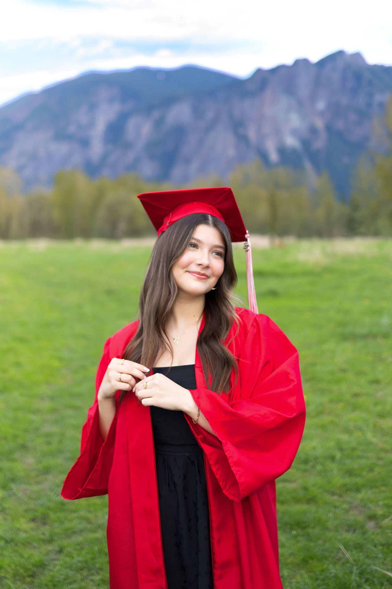 Graduate in bright red academic regalia poses outdoors against mountain backdrop and green field on graduation day.
