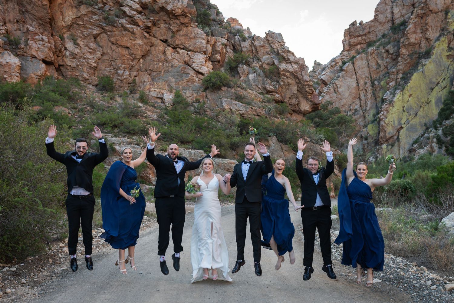 Wedding party jumping joyfully on a dirt path with rocky canyon backdrop, bride in white gown and groomsmen in tuxedos.