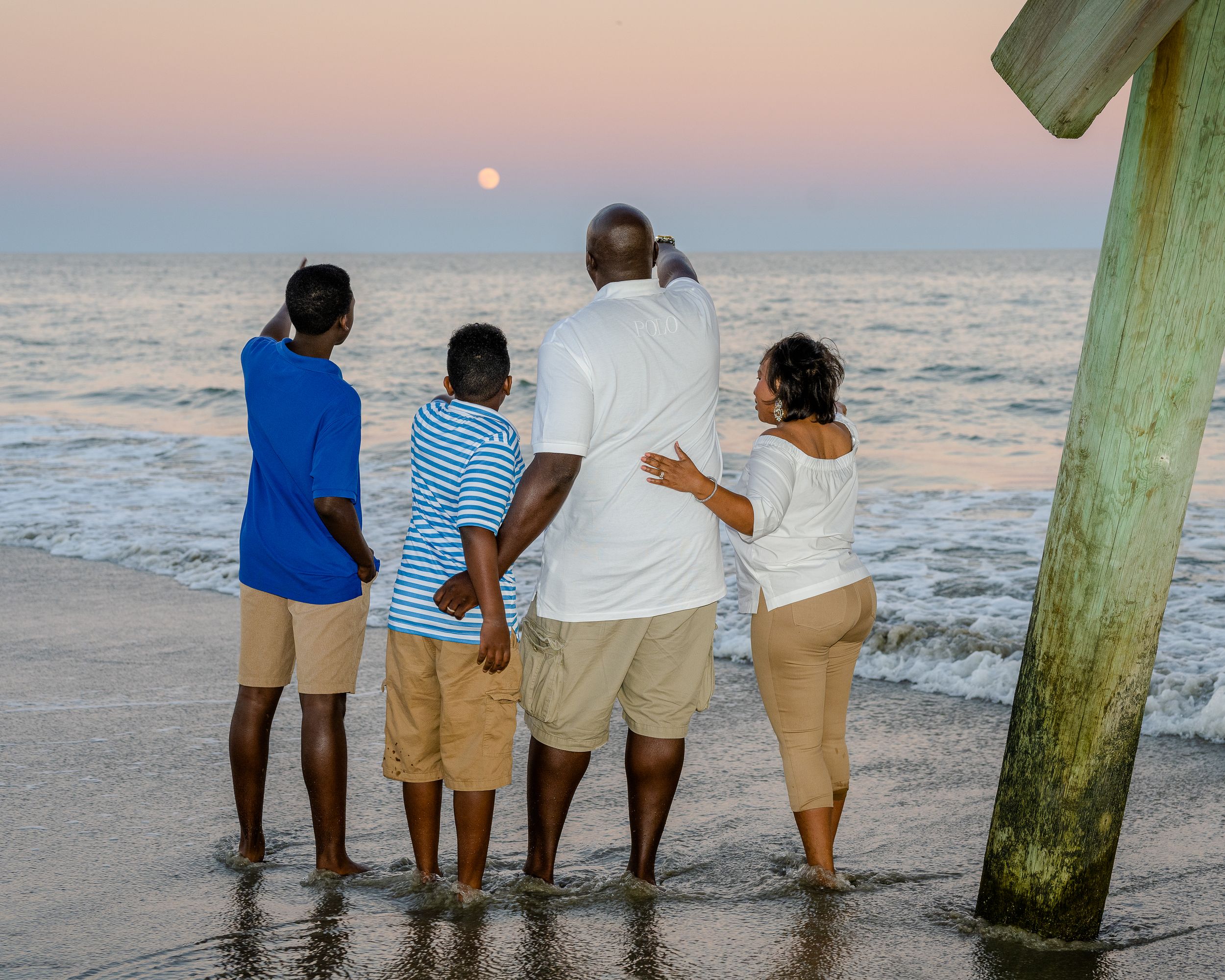A group of people sit together on a wooden pier watching a pink sunset over the ocean.