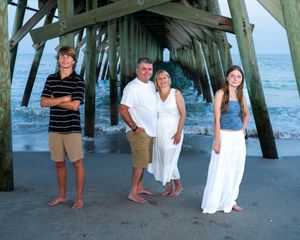 Family group posing for a beach photo under a wooden pier with ocean waves and blue water in the background.