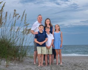Family members stand together on a sandy beach with sea oats and ocean waves in the background.