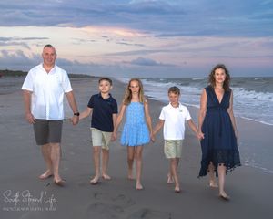 A family of five walking together on a sandy beach at sunset with ocean waves and cloudy skies in the background.