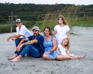 A group of people sitting casually on a sandy beach with grasses and greenery in the background.