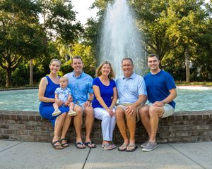 A group in coordinating blue outfits sits together on a brick wall in front of a fountain on a sunny day.
