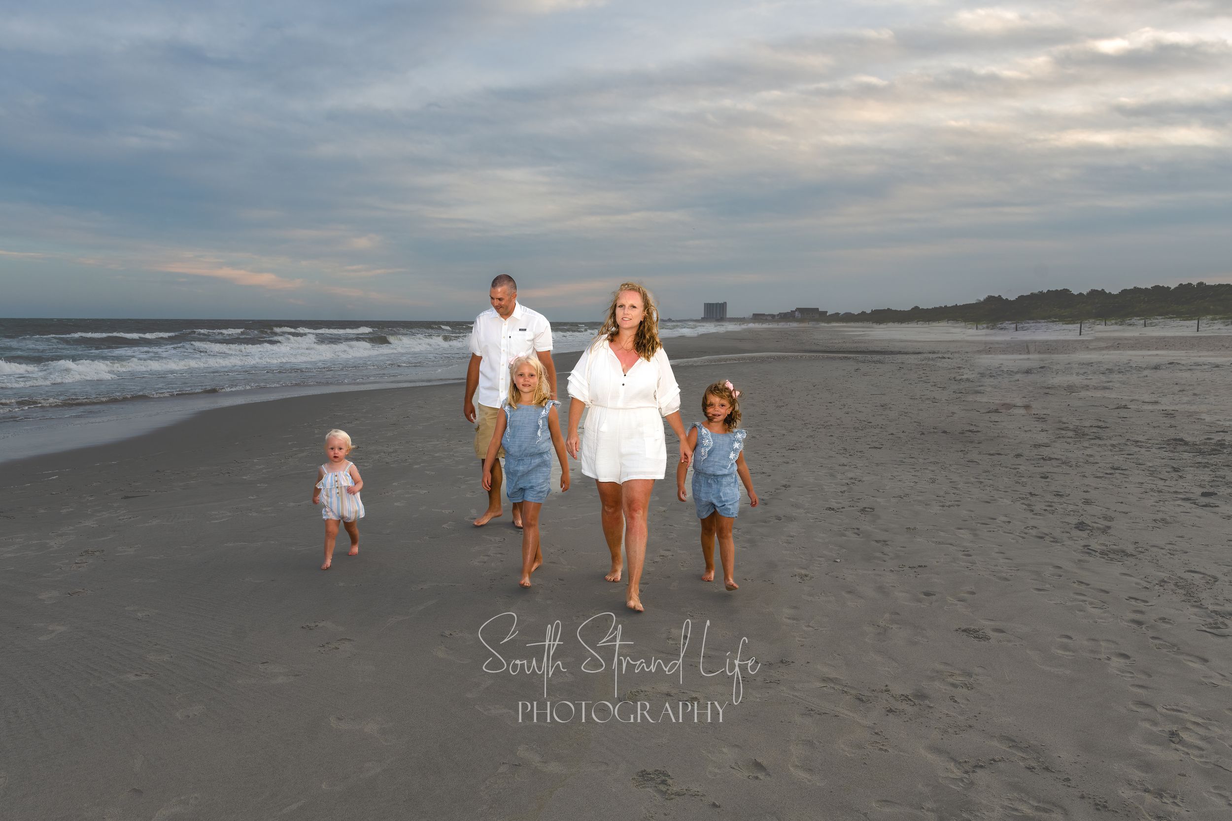 Family walking along a sandy beach at sunset with cloudy skies in the background.