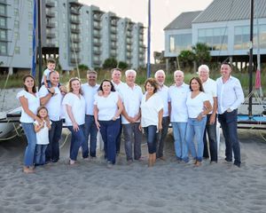 A group of people wearing white and blue clothing pose together on a beach at sunset.