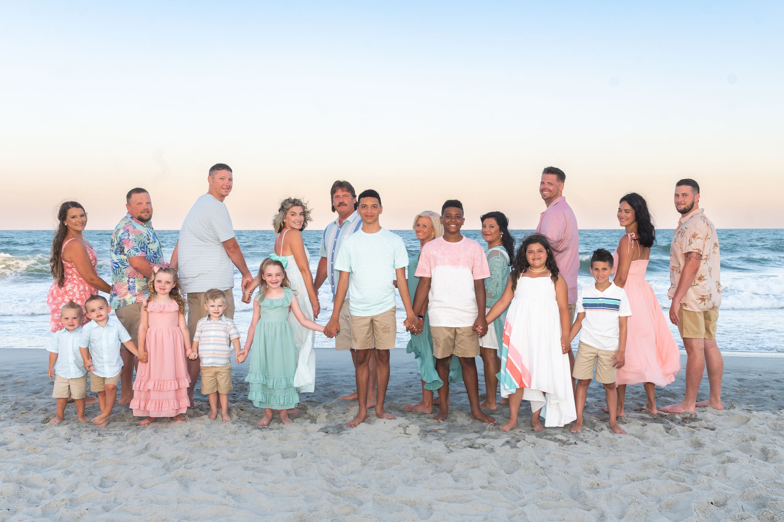 Large family group wearing coordinated pastel outfits poses together on beach at sunset.