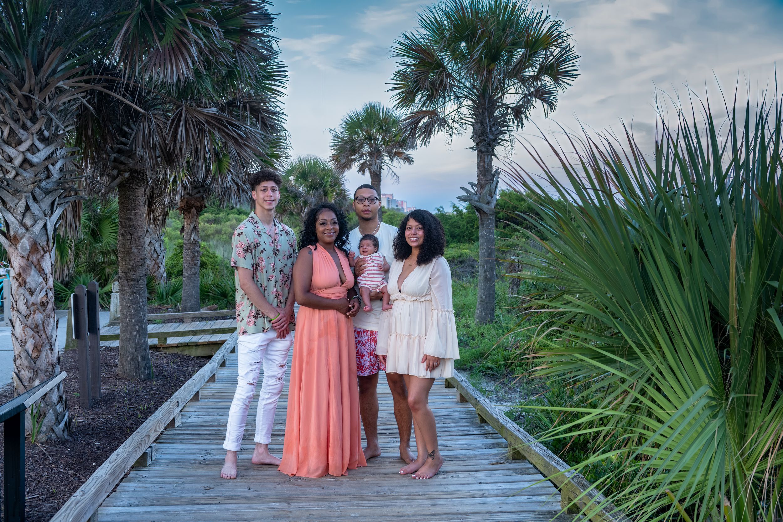 People walking along a scenic boardwalk path lined with palm trees and tropical vegetation at sunset.