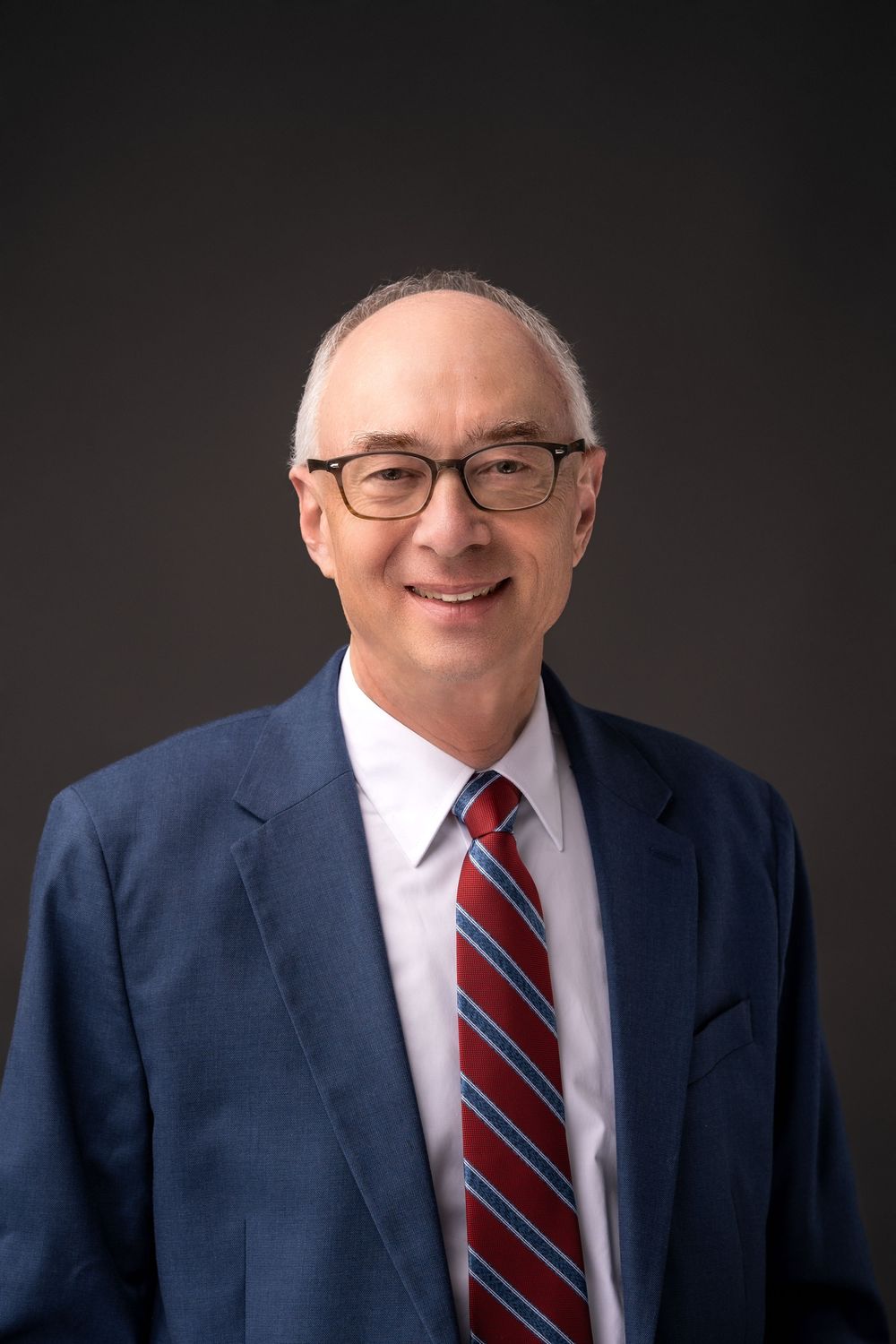Executive portrait in navy suit and striped tie against dark background.