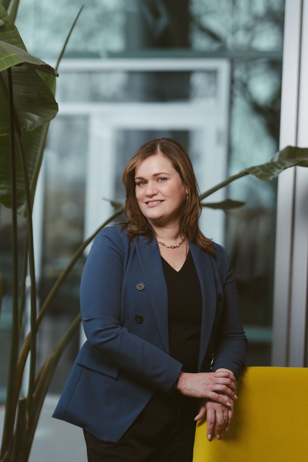 Professional seated pose near window with tropical plants wearing navy blazer.