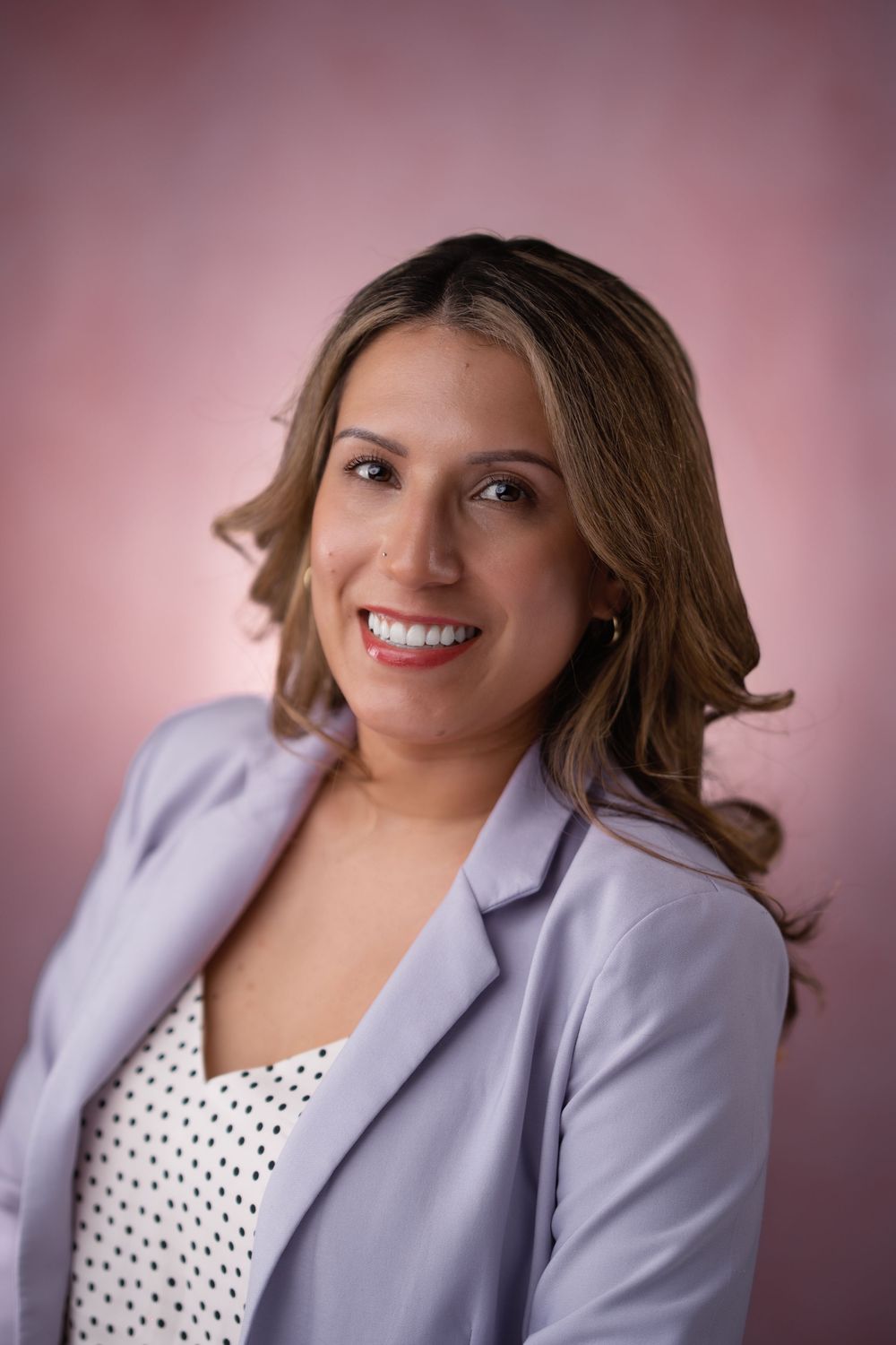 Professional headshot of woman in lavender blazer and polka dot top against pink backdrop.