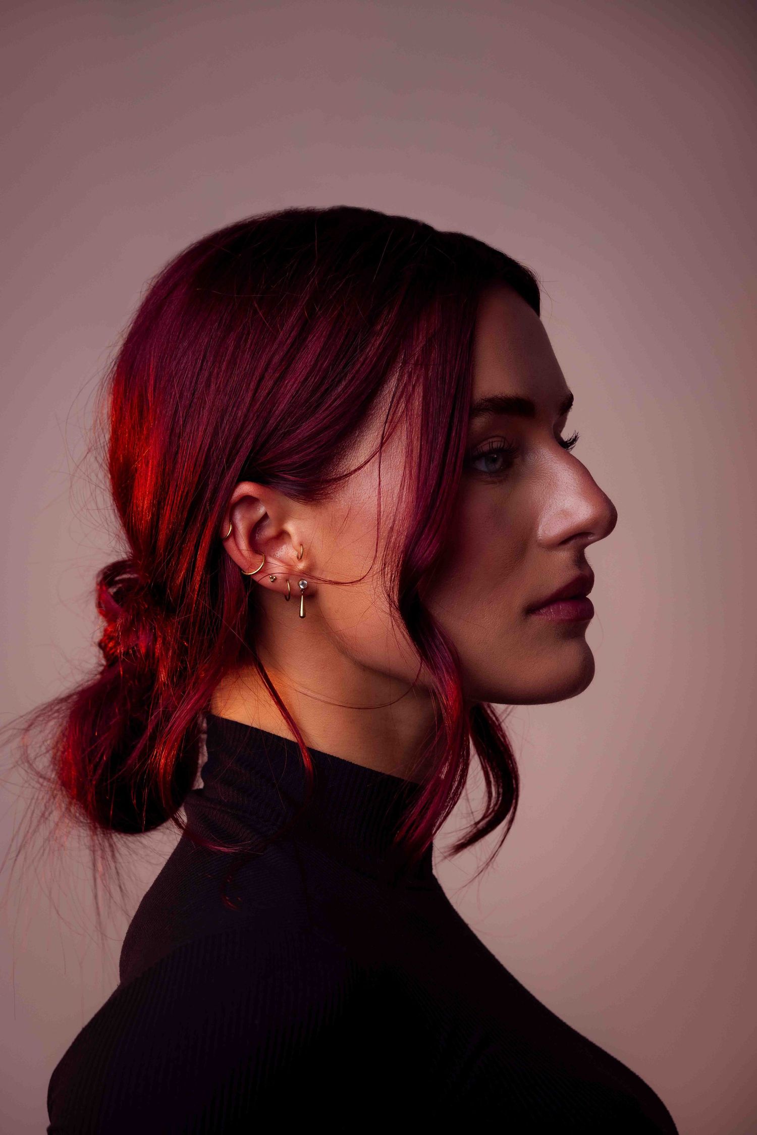 A modeling portrait of a woman with vibrant red hair styled in an updo wearing a black top against a soft pink background.