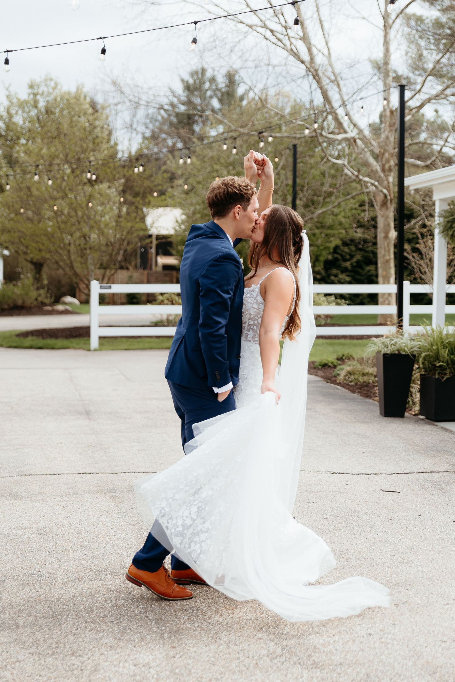 A couple in wedding attire performs a romantic dip pose on a gravel path surrounded by trees and string lights.