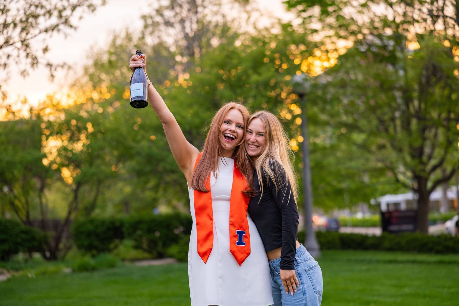 Graduating girl student holding a bottle of champagne while hugging her friend at UIUC