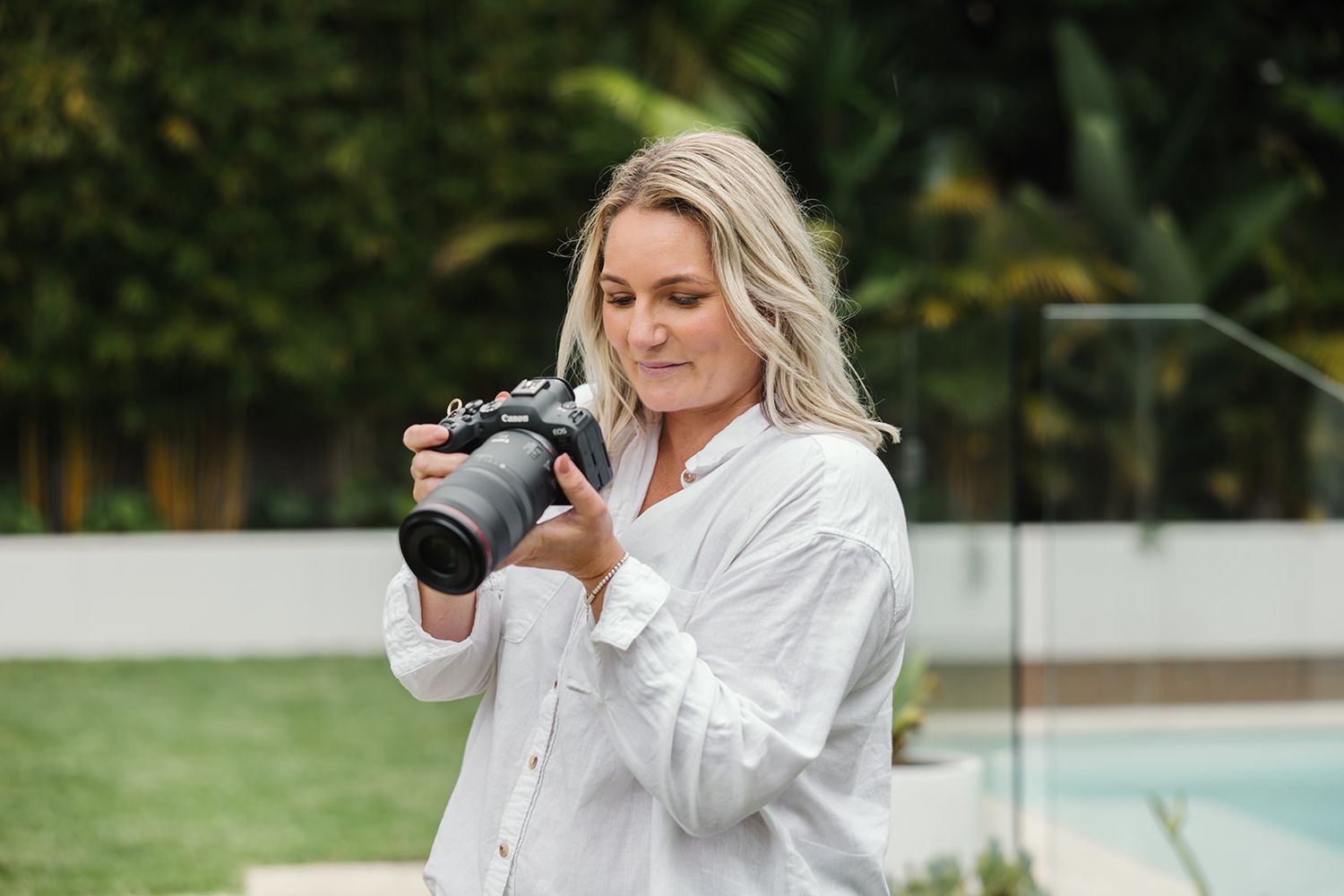 A photographer in a white button-down shirt uses a professional DSLR camera outdoors near a pool.