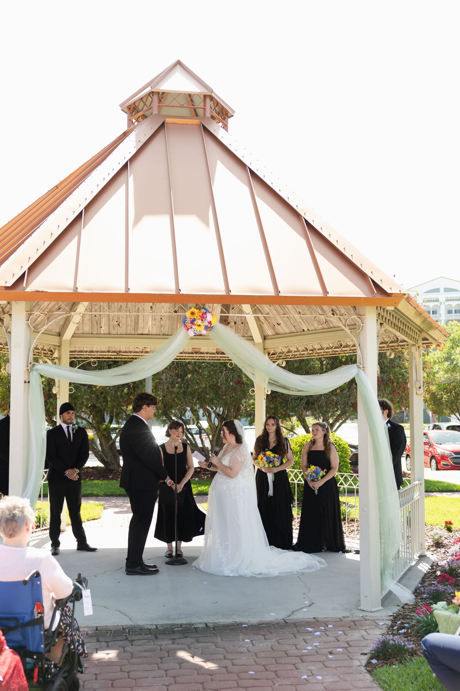 Outdoor wedding ceremony under a white gazebo with bride, groom, officiant, and bridesmaids in black dresses.