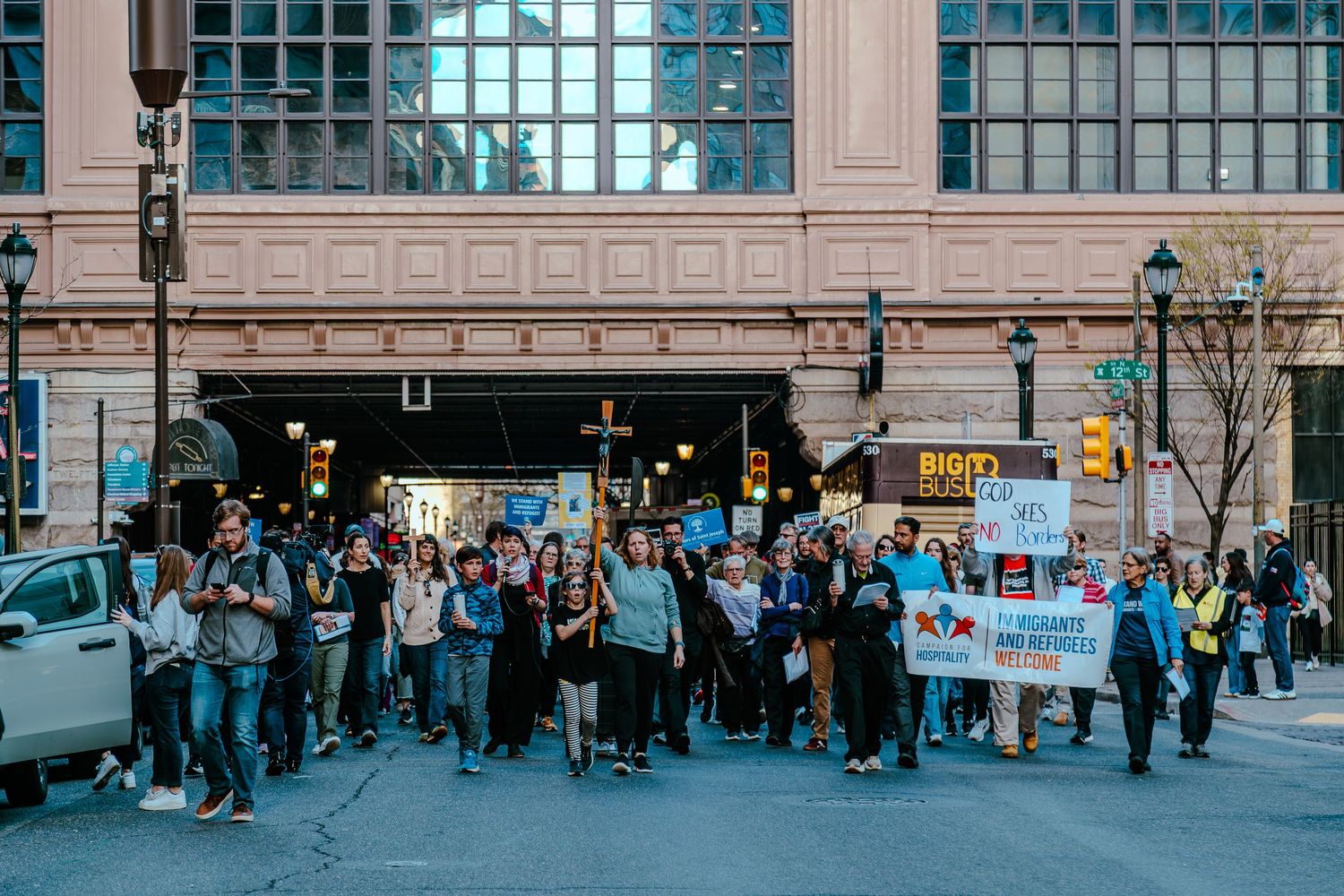 Large group of protesters marching through urban streets holding signs and banners during a public demonstration.