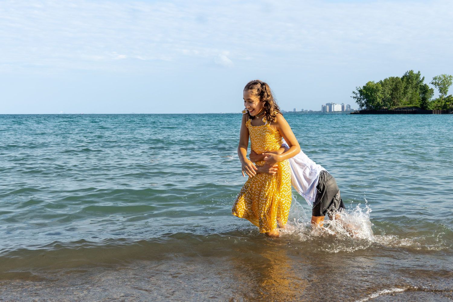 A woman in a yellow dress walks through the shallow waters of a beach with a city skyline in the background.