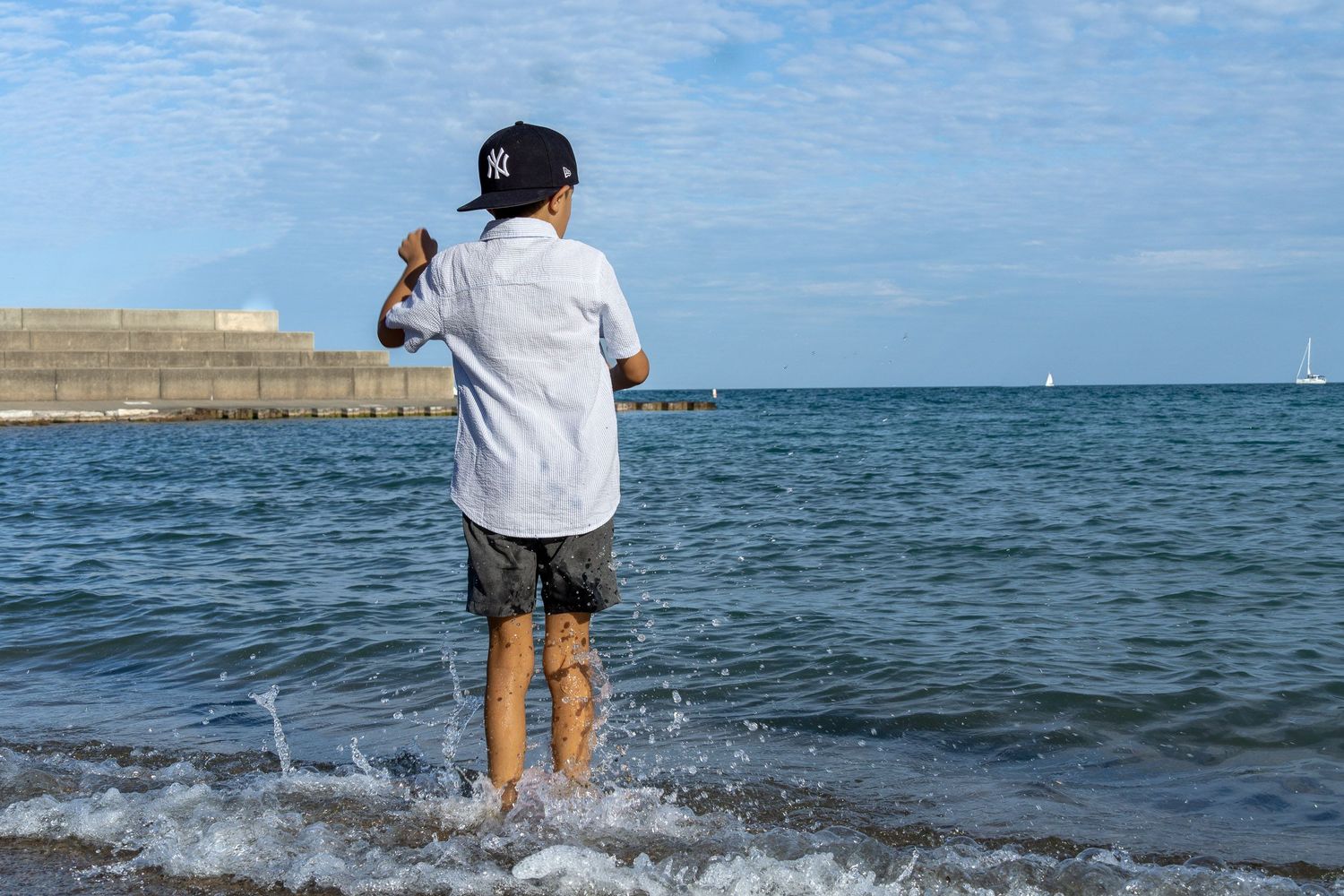 A person wearing a white shirt and shorts stands in the shallow water of a lake or ocean, with a blue sky and horizon visible.