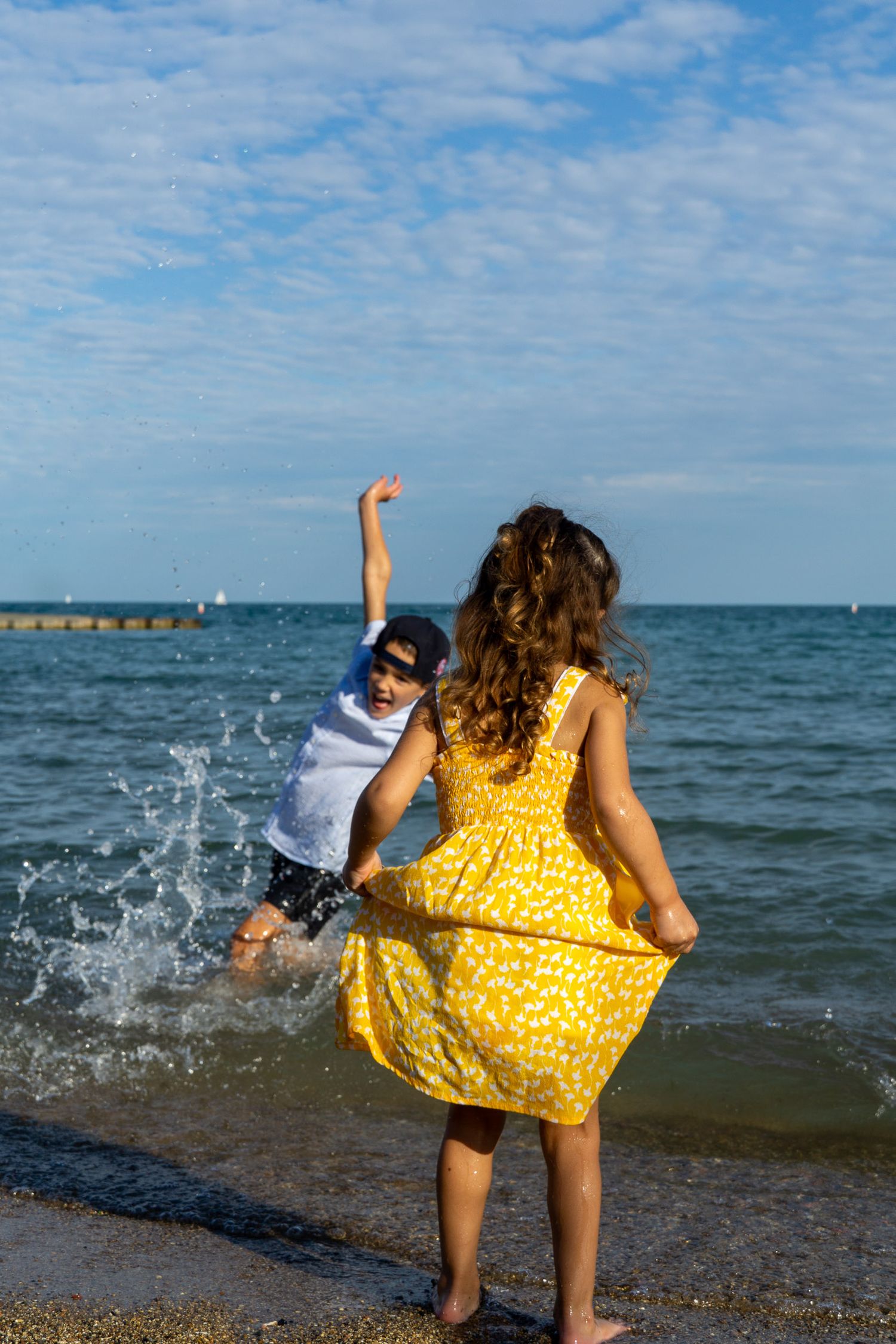 Kids in a yellow dress stands in the waves of a beach, splashing water with a young child beside her.