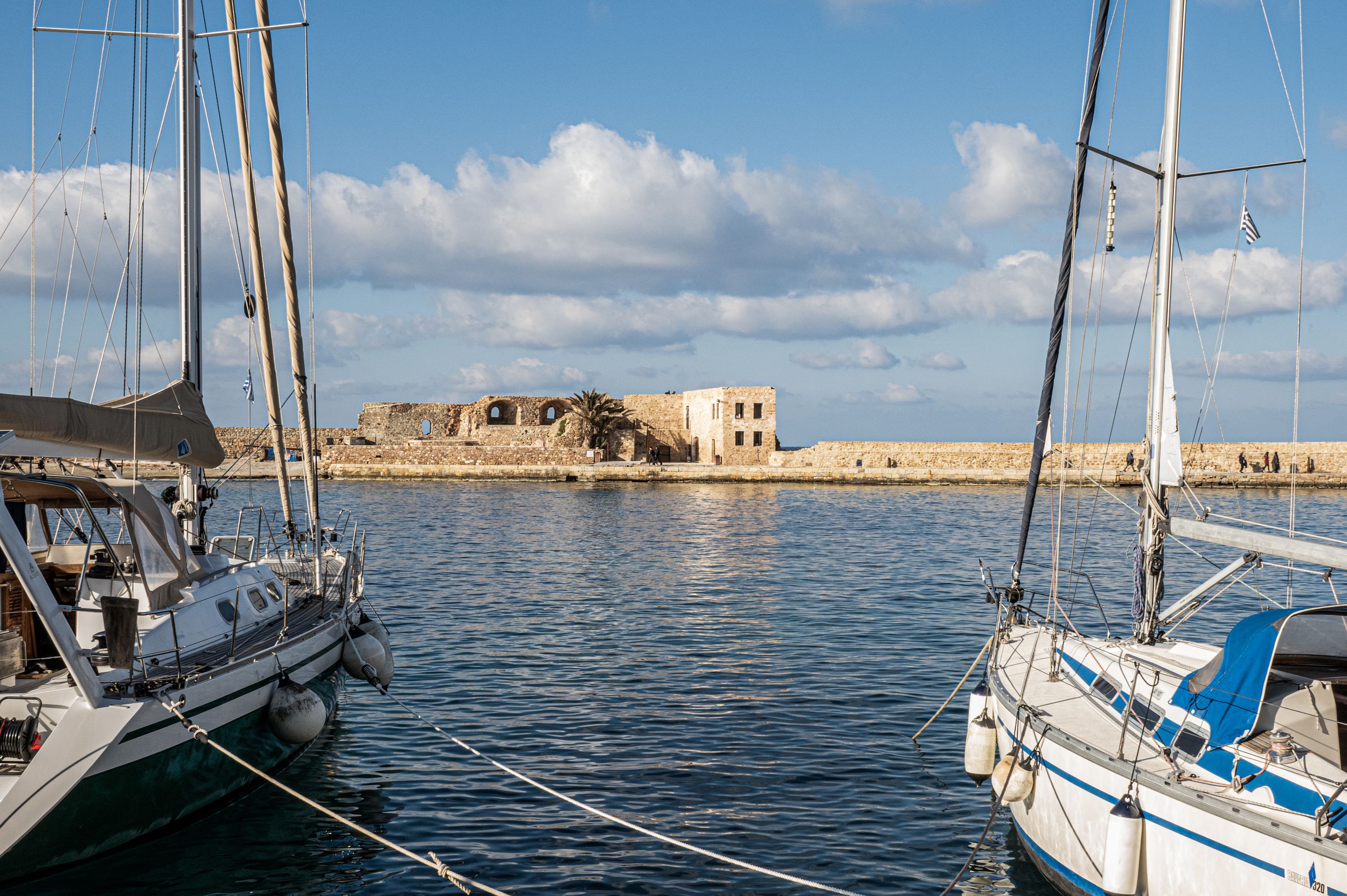 A series of sailing boats anchored in a calm blue harbor with coastal buildings visible in the background.
