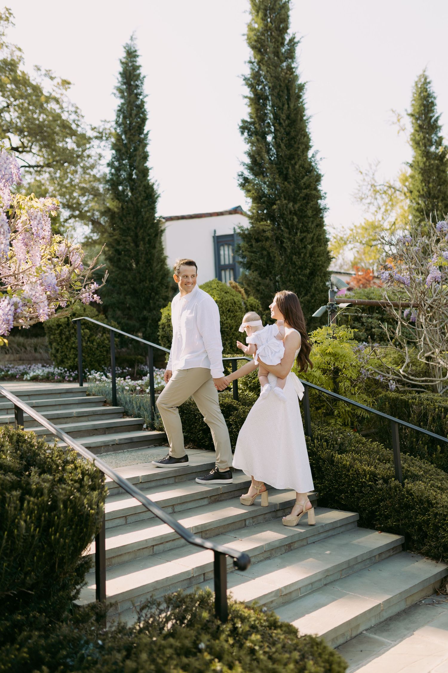 A couple in white outfits walking up outdoor stone steps surrounded by cypress trees and garden landscaping.