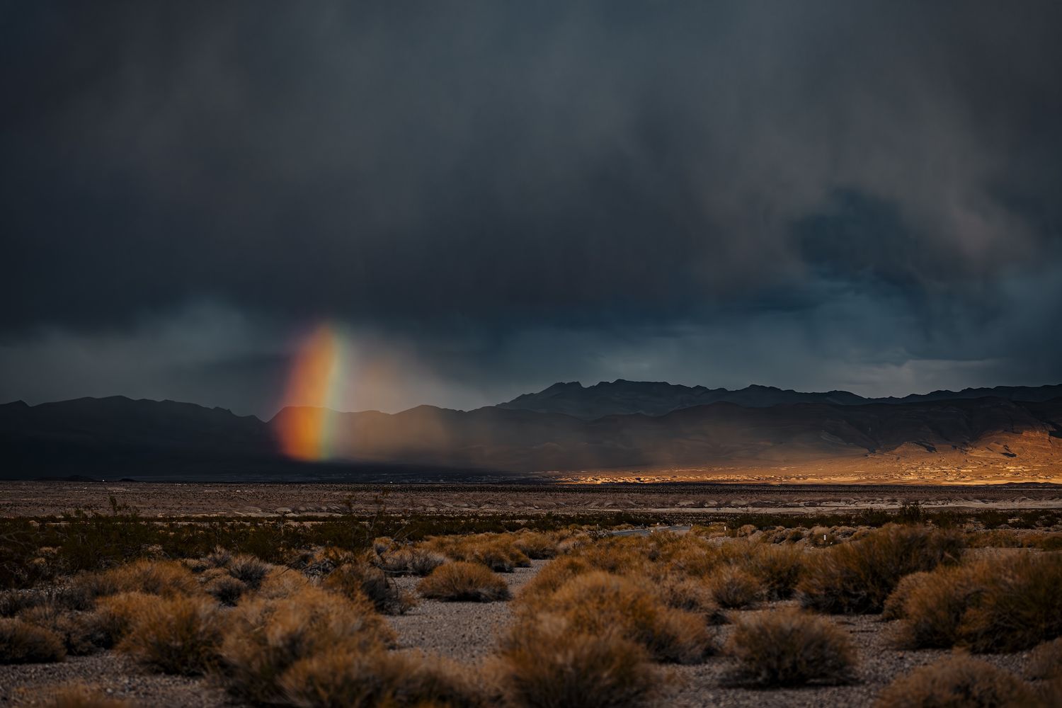 A vibrant rainbow emerges over a dramatic desert landscape with stormy skies and mountains in the background.