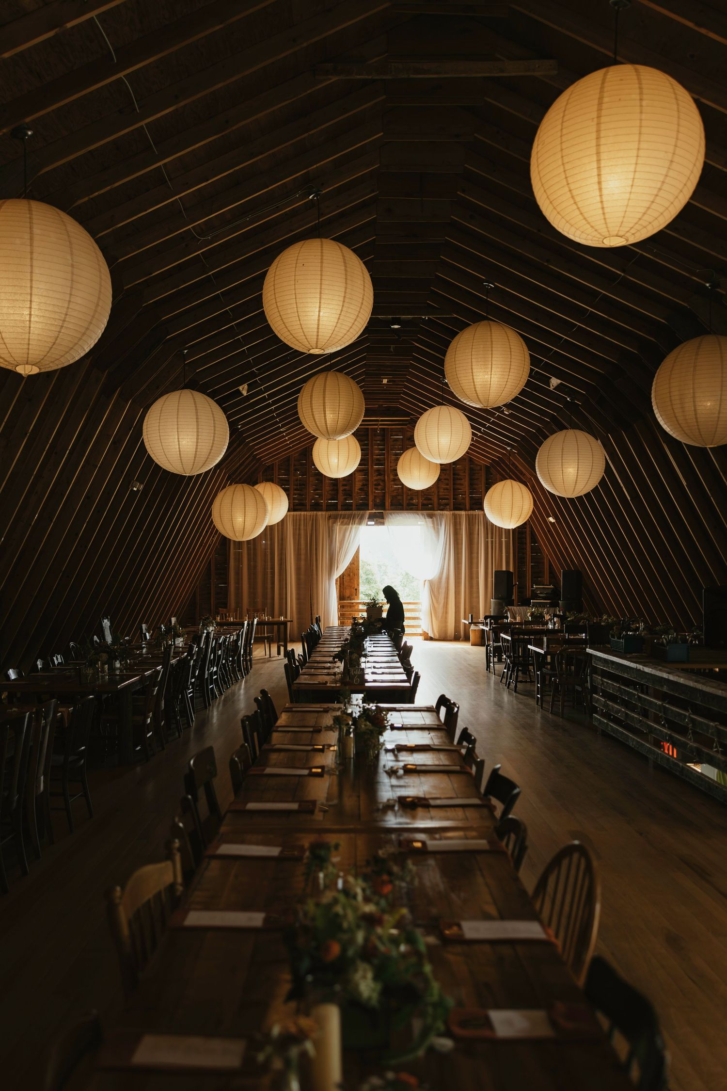A row of glowing paper lanterns illuminate a dark wooden interior space with tables and chairs arranged in rows.
