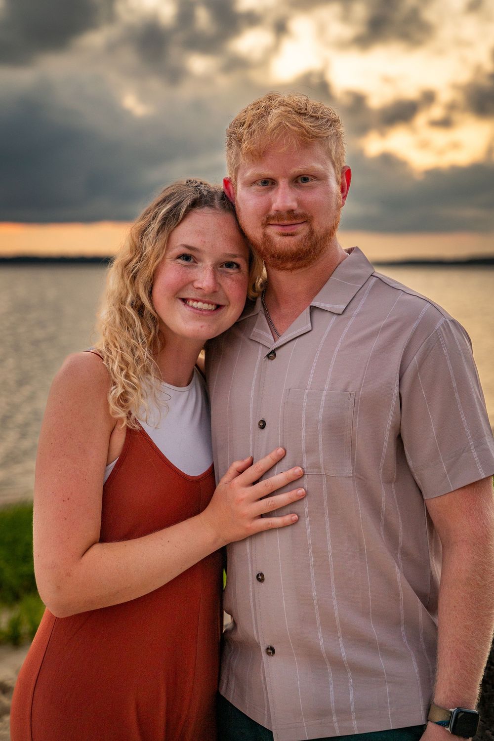 A couple poses together at sunset by the water wearing rust-colored and gray clothing.