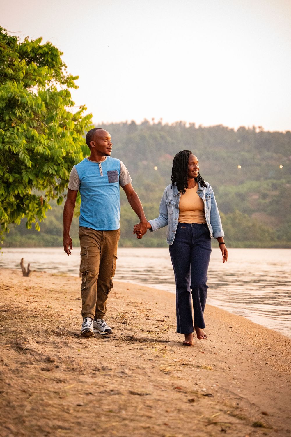 A couple walks hand in hand along a scenic beach at sunset with hills and trees in the background.