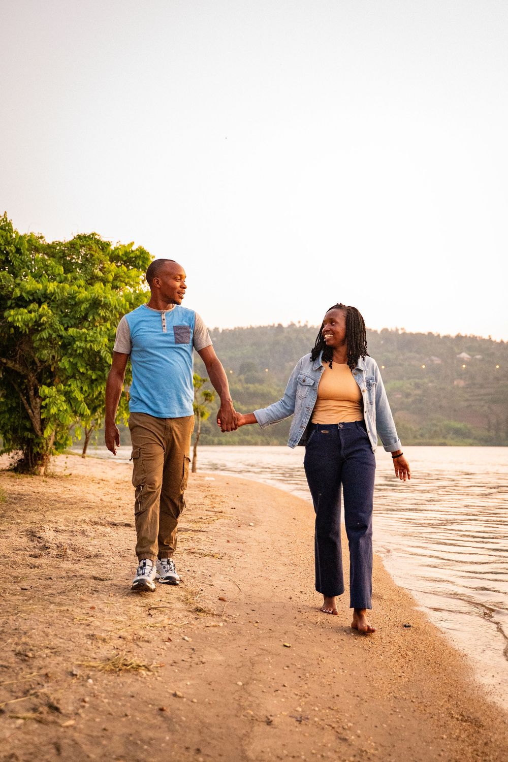 A couple enjoys a romantic walk along a sandy beach shoreline during golden hour with hills in the background.