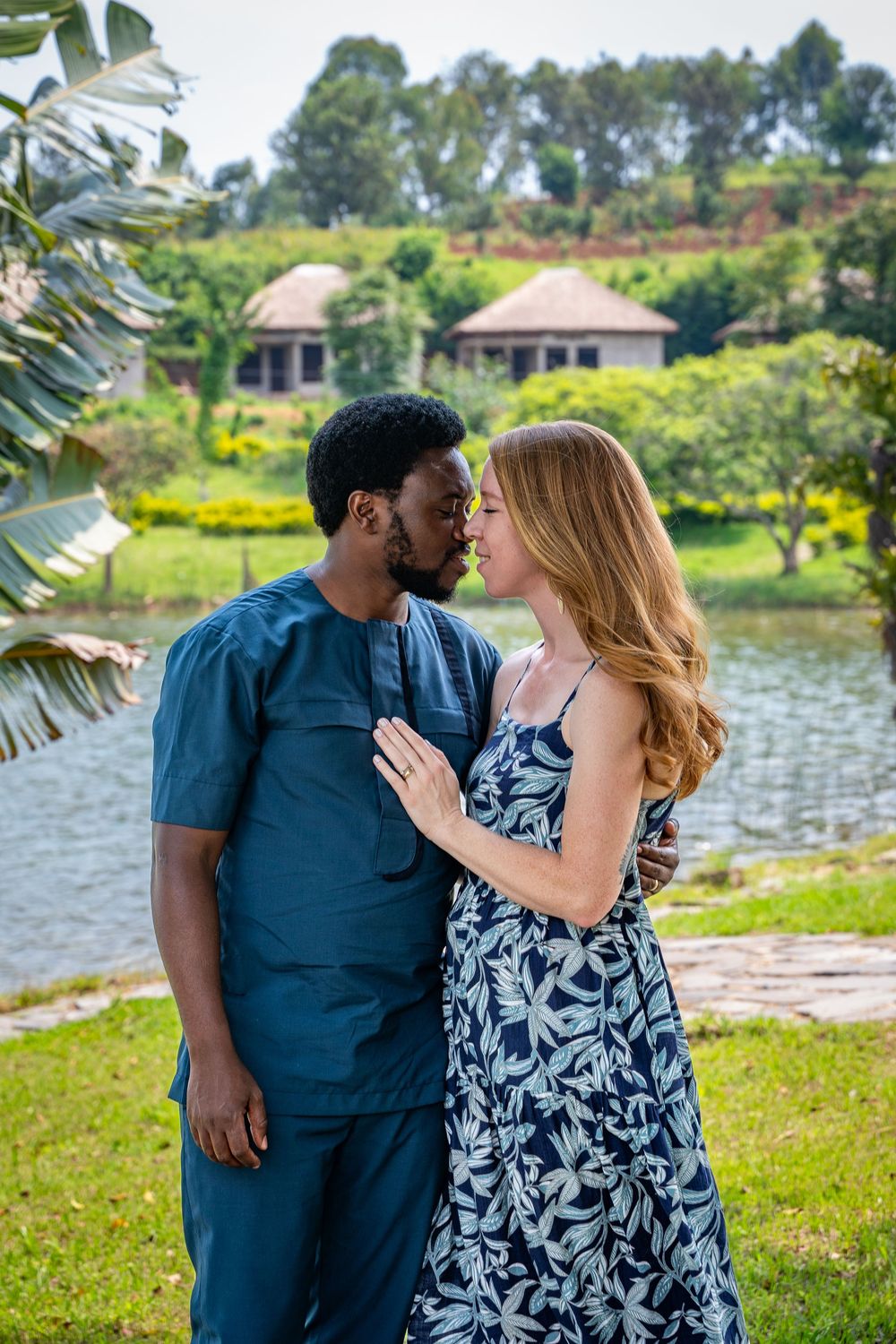 A couple shares romantic moments by a lakeside surrounded by lush greenery and palm trees.
