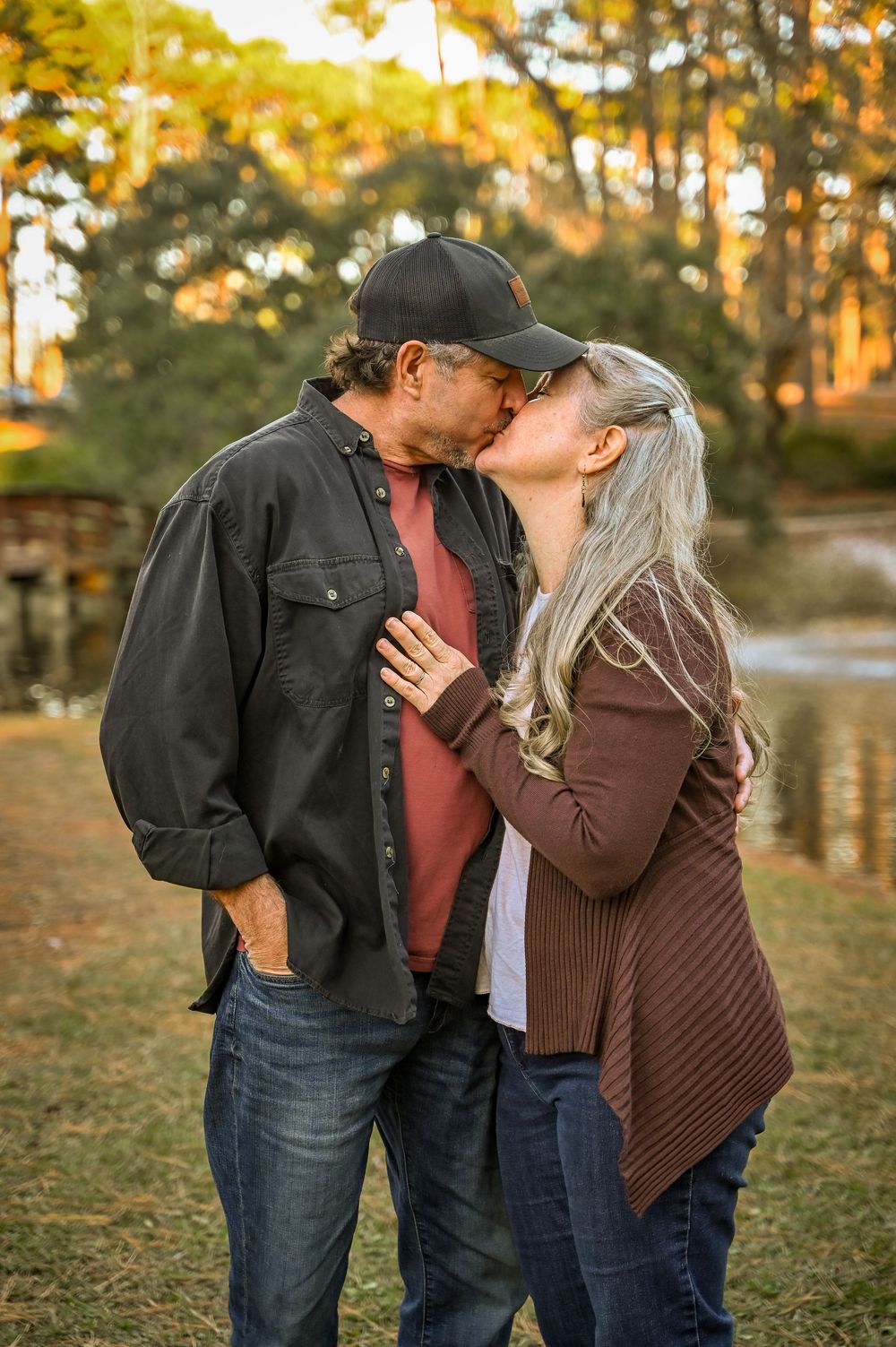A couple shares a romantic kiss by a lake during golden hour sunset in autumn, surrounded by fall foliage.