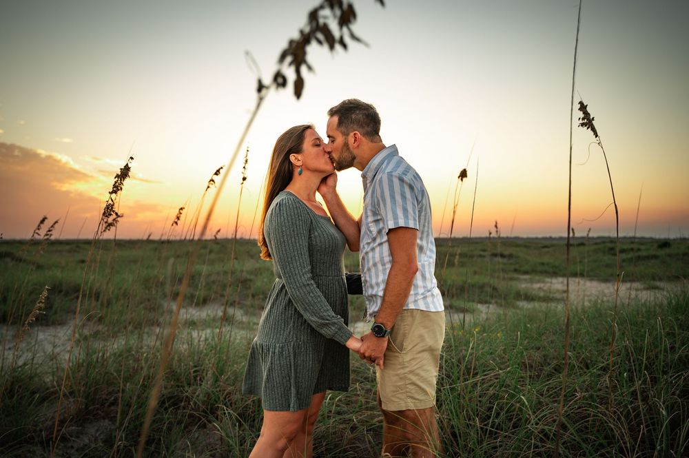 Romantic sunset photoshoot with couple silhouetted against orange sky and tall grass.