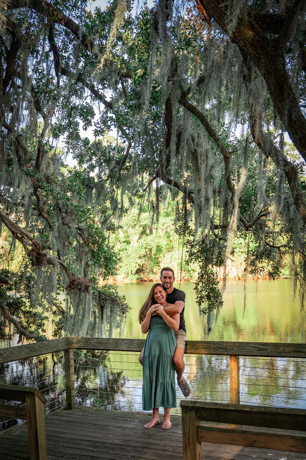 Couple embracing on wooden boardwalk overlooking serene lake with Spanish moss draped trees.