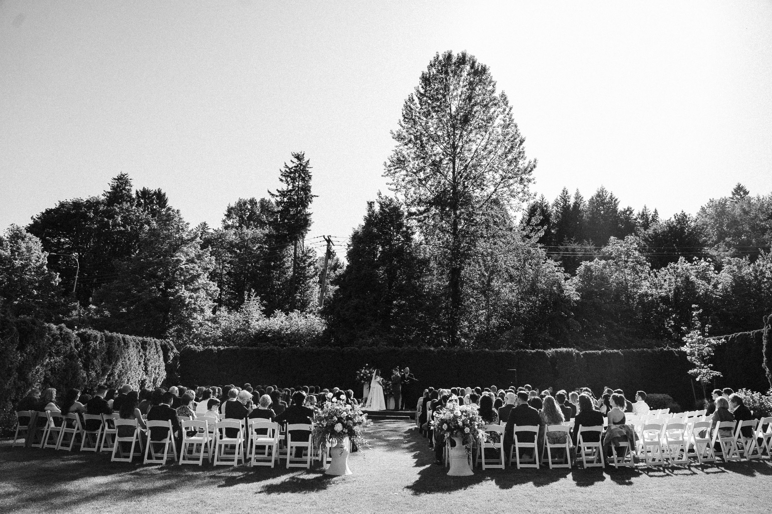 Black and white outdoor wedding ceremony setup with rows of wooden chairs facing a garden backdrop.