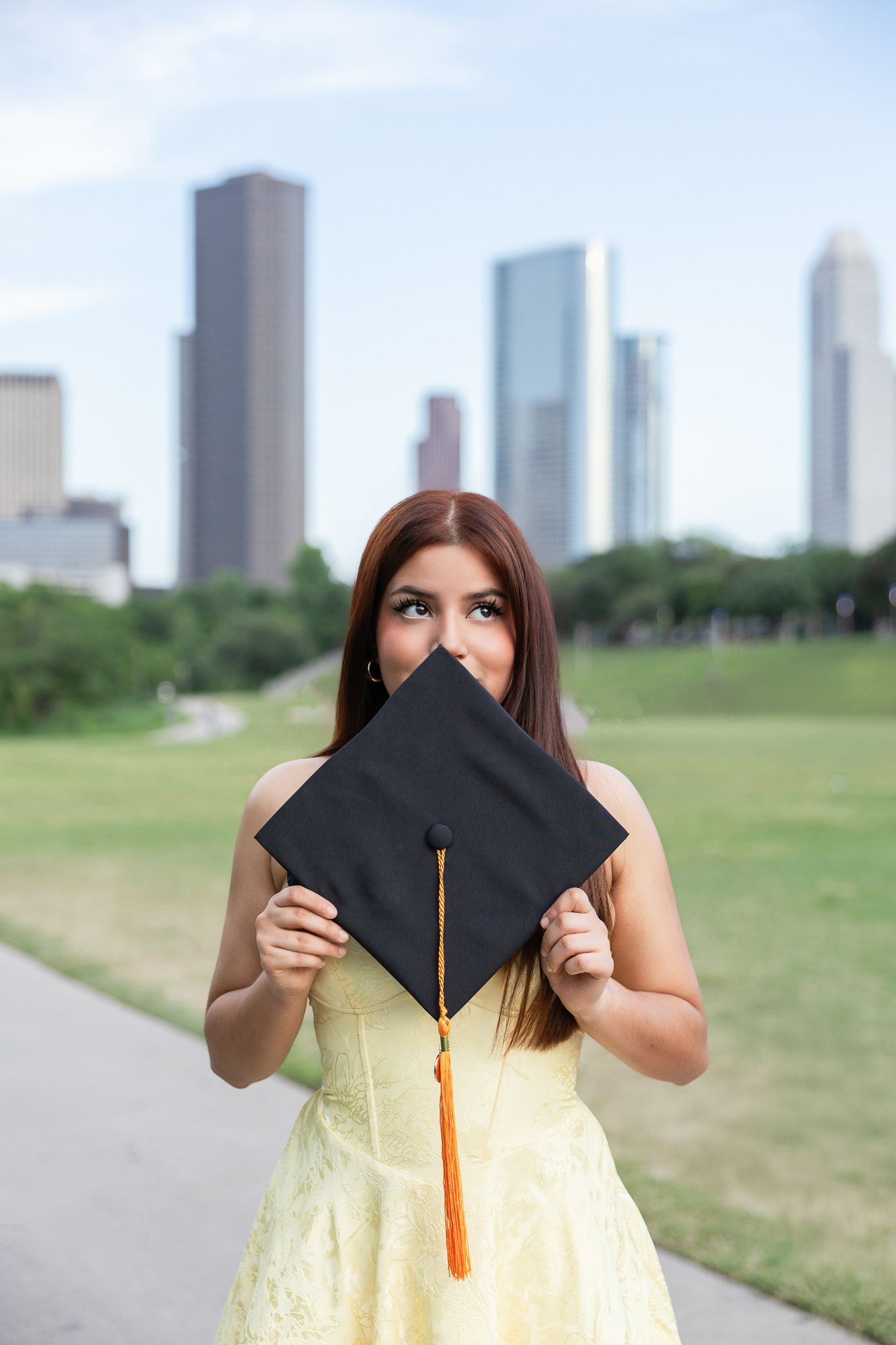 Graduate holding up black cap with orange tassel in front of Houston skyline in yellow dress.